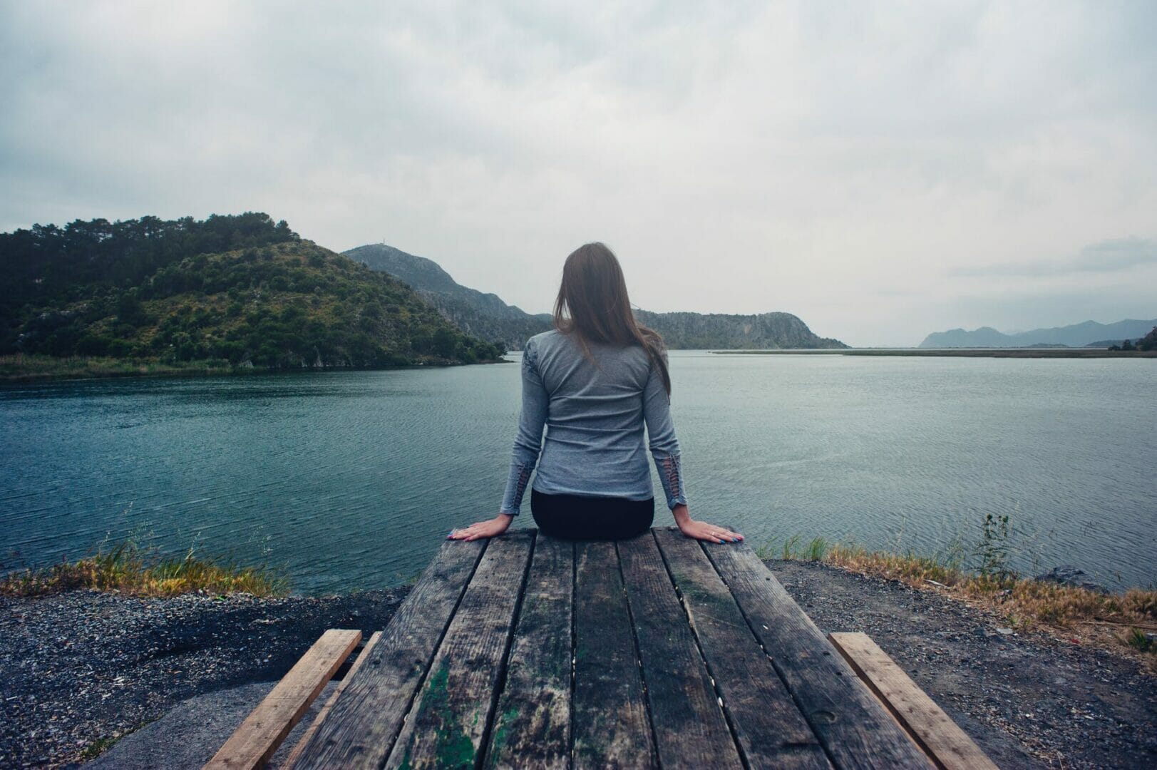 woman wearing gray long sleeved shirt and black black bottoms outfit sitting on gray wooden picnic table facing towards calm body of water at daytime - Vacation Instagram Captions 